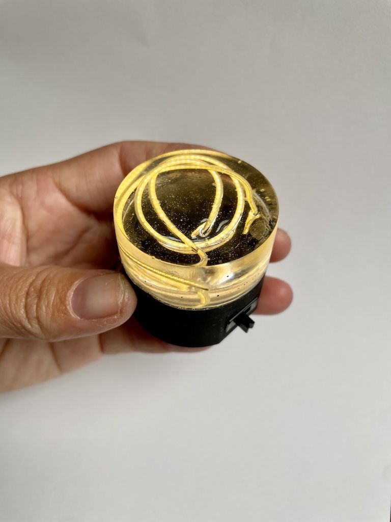 A small cylindrical puck in a hand. The top half is clear resin with a lit LED filament in loops inside, the bottom half is black 3D print.