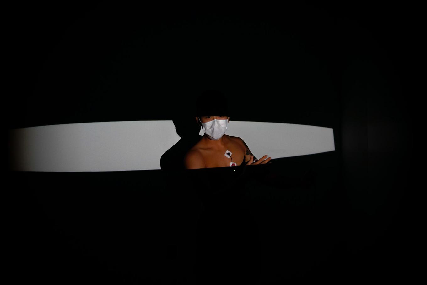 One slightly rounded slit of light, horizontal across the frame, carves space out of the darkness. In the center of it is revealed the upper torso and lower face (masked) of a person (Spencer).