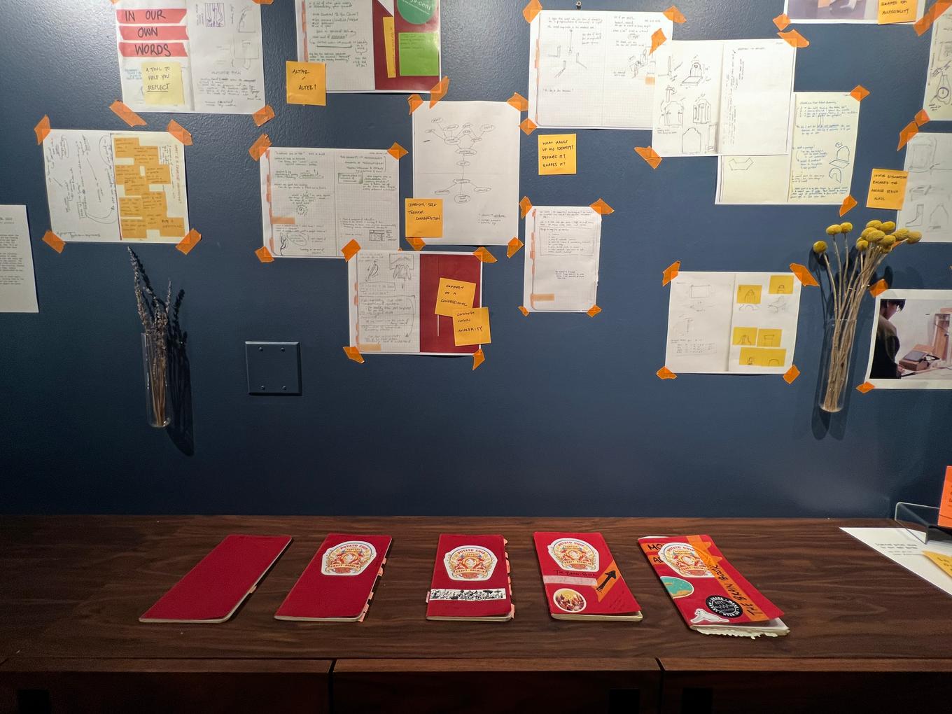 Front-facing image of wall, including many pages of sketches, writing, and ideation taped with orange tape. On a shelf at the bottom of the image is 5 notebooks.